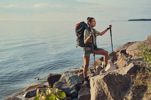 Göl kenarındaki taşlara tırmanmak için İskandinav bastonları kullanan kadın. — Stok fotoğraf