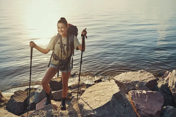 Happy hiker smiling at camera while standing on lakeside rocks — Stock Photo, Image