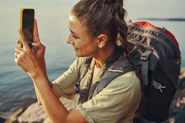 Sırt çantalı akıllı telefondan göl manzarasının fotoğrafını çekiyor. — Stok fotoğraf