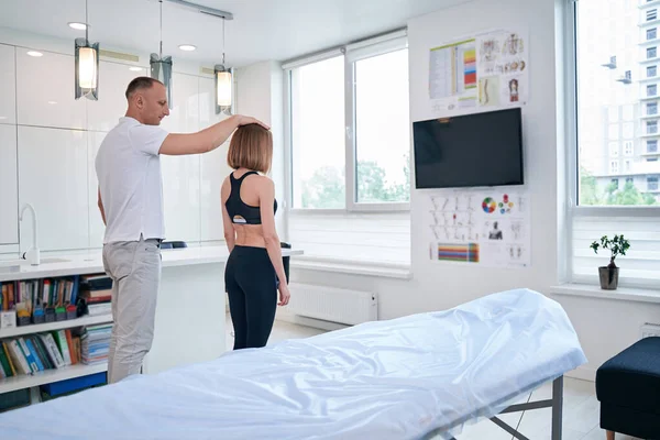 Young Caucasian man osteopath examining back of charming redheaded woman in black sportswear — Stock Photo, Image