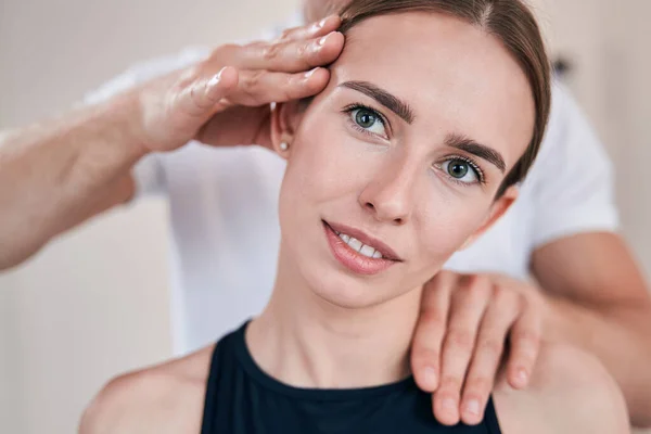 Vacker kvinna poserar vid fotokameran medan oigenkännlig massage terapeut undersöker hennes hals — Stockfoto