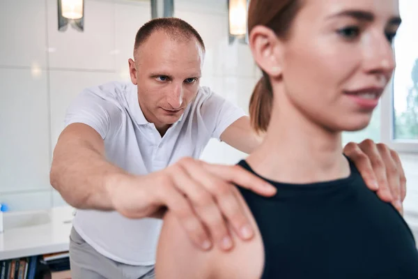 Handsome neat physiotherapist examining shoulder of his woman client in medical office — Stock Photo, Image