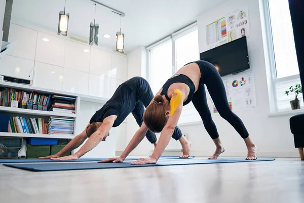 Beautiful elegant lady and neat male practicing down ward dog yoga pose in modern studio — Stock Photo, Image