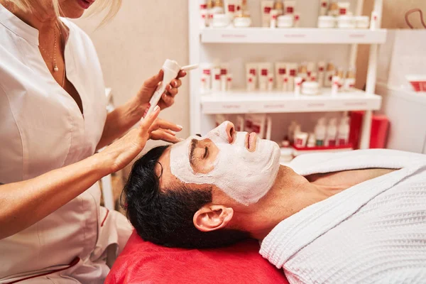 Массажный терапевт наносит увлажняющую маску на лицо клиента — стоковое фото