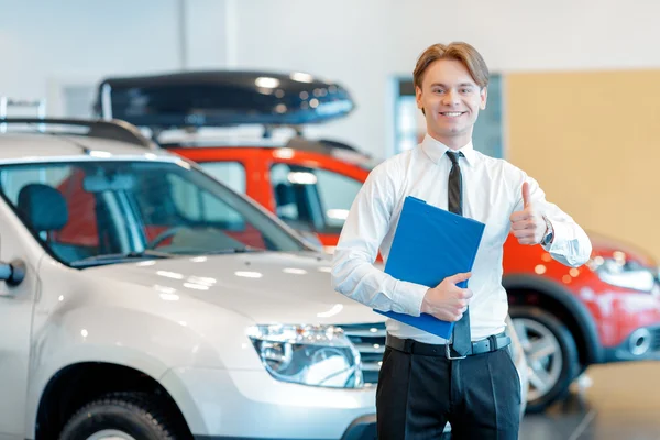 Jovem vendedor sorridente com prancheta na concessionária de carros — Fotografia de Stock