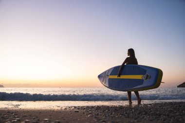 Genç beyaz kadın gün batımında sakin plaj boyunca raket taşıyor. Sakin okyanus ve ılık renkler huzurlu bir atmosfer yaratır, yansıma ve sükuneti davet eder..