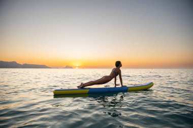 Sakin bir kadın güneş doğarken okyanusta bir sörf tahtasında yoga yapıyor. Huzurlu ortam ve huzurlu ruh hali bir huzur ve farkındalık hissi uyandırıyor..