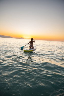 Sakin bir kadın, sakin gündoğumunda açık okyanusta kürek çekmeyi sever. Yumuşak ışık huzuru ve doğayla bağlantıyı yansıtan barışçıl bir atmosfer yaratır..
