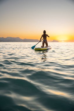 Açık tenli genç bir kadın gün batımında okyanusta sörf yapmayı sever. Sakin ortam ve sakin sular barışçıl bir atmosfer yaratır..