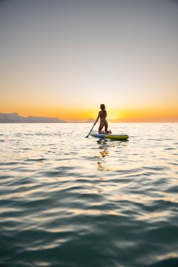 Genç kadın gün batımında sakin okyanusun tadını çıkararak kürek tahtasında diz çöker. Sakin atmosfer, sakin duyguları ve huzurlu açık hava deneyimini yansıtır..