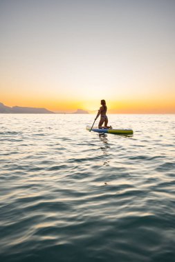 Genç bir kadın güzel günbatımında sakin okyanusta kürek çekmeyi sever. Huzurlu atmosfer ve huzurlu ifade, doğayla olan huzuru ve bağlantıyı yansıtır..