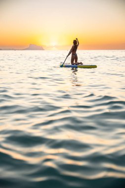 Genç bir kadın çarpıcı bir günbatımında okyanusta kürek çekmeyi seviyor. Huzurlu atmosfer, onun sakin ifadesini ve doğayla olan bağlantısını vurgulayan barışçıl bir sahne yaratır..