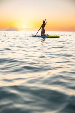 Güzel bir gündoğumu sırasında genç bir kadın sakin okyanus sularında zarif bir şekilde kürek çekiyor. Barışçıl atmosfer huzuru ve doğayla bağlantıyı çağrıştırıyor..