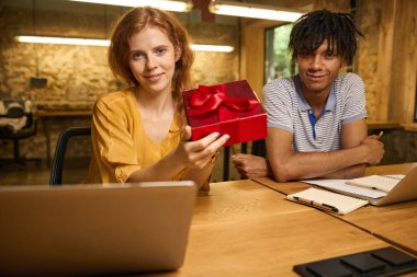 Sarı bluzlu bir kadın kırmızı bir hediye kutusunu dikkatlice açarken çizgili gömlekli bir adam dikkatle izliyor.