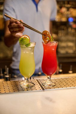 Barın arkasında barmen, elinde dekoratif süslerle iki canlı kokteyl tutarken gülümsüyor.