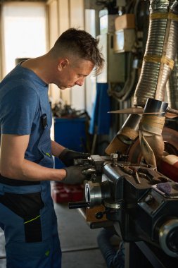 Mavi üniformalı tamirci atölyeden kaldırılan bir araba üzerinde çalışıyor.