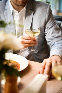 Restoranda yemek yerken elinde bir bardak beyaz şarap tutan takım elbiseli bir adamın yakın çekimi.