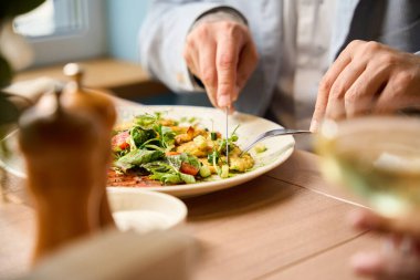 İki kişi yemek yiyor, biri yemek masasında salata yemek için çatal ve bıçak kullanıyor.