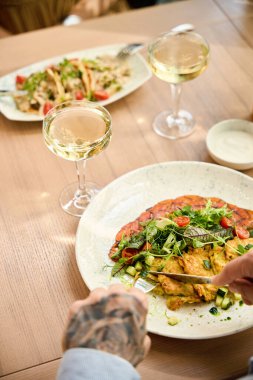 Arkadaşı karşısında otururken kolunda dövme olan adam çatal bıçak kullanıyor.