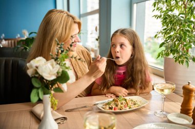 Kadın küçük kızına bir restoranda masada otururken salata yediriyor.