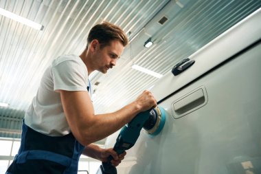 Atölyede arabasının yanında elinde cilalama makinesi olan üniformalı genç bir adam. Araba temizleme kavramı