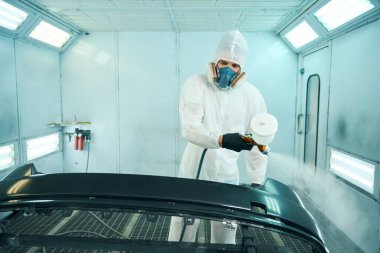 Koruyucu üniformalı bir adam arabanın parçalarını özel boya odasına boyuyor. Araba servisi konsepti