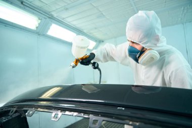 Koruyucu takım elbiseli tamirci boya odasındaki vücut elemanlarını boyuyor. Araba servisi konsepti