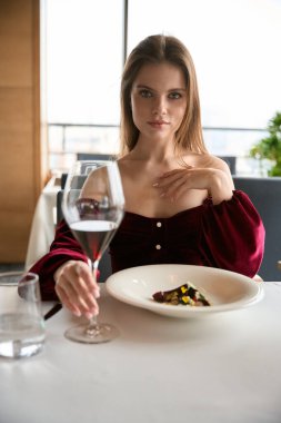 Restoranda oturmuş kırmızı şarap içip kameraya poz veren güzel ve genç bir bayan. Yaşam tarzı, hizmet kavramı