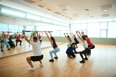 Sportif erkekler ve kadınlar jimnastik sopaları tutan kadın antrenörle çömelme hareketleri yapıyorlar. Fitness, spor, eğitim, spor salonu ve yaşam tarzı konsepti