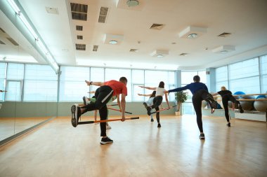Bir grup insan tek ayak üzerinde durup jimnastik sopalarını tutuyor. Fitness, spor, eğitim, spor salonu ve yaşam tarzı konsepti