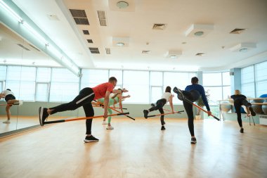 Bir grup insan spor salonunda jimnastik sopasıyla tek ayak üzerinde denge kurarken egzersiz yapıyor. Fitness, spor, eğitim, spor salonu ve yaşam tarzı konsepti