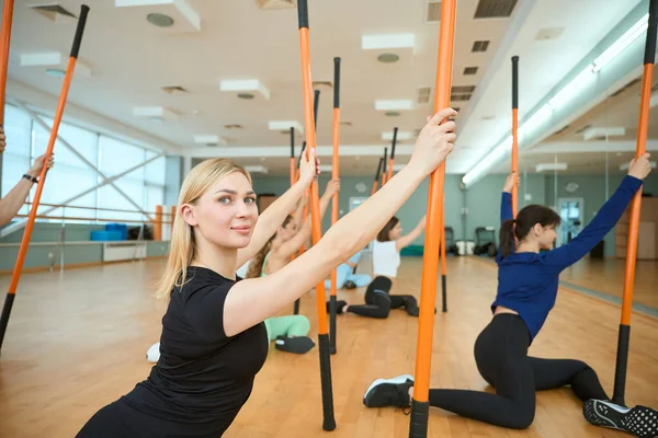 Jimnastik çubuğu kullanan mutlu genç bayan spor salonunda insanlarla esneme hareketleri yaparken yerde oturuyor. Fitness, spor, eğitim, spor salonu ve yaşam tarzı konsepti
