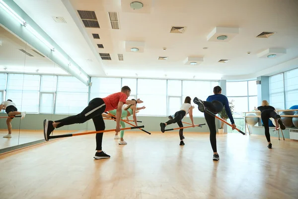 Bir grup insan spor salonunda jimnastik sopasıyla tek ayak üzerinde denge kurarken egzersiz yapıyor. Fitness, spor, eğitim, spor salonu ve yaşam tarzı konsepti