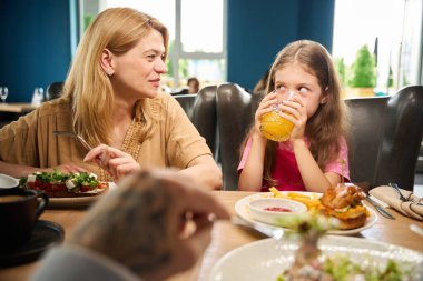 Genç kız meyve suyundan bir yudum alırken annesi gülümseyerek seyrediyor, sakin bir anı paylaşıyor.