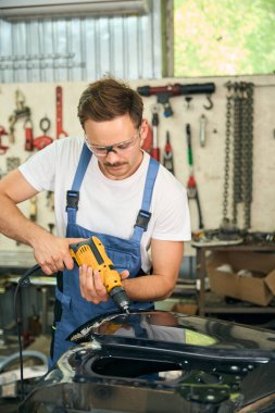 Koruyucu gözlüklü bir adam oto tamirhanesinde elektrikli tornavidayla araç parçalarını tamir ediyor. Araba tamir hizmeti kavramı