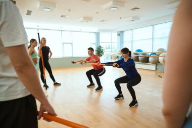 Spor salonunda jimnastik sopası olan insanlara nasıl çömelmesi gerektiğini anlatan kadın koç. Fitness, spor, eğitim, spor salonu ve yaşam tarzı konsepti
