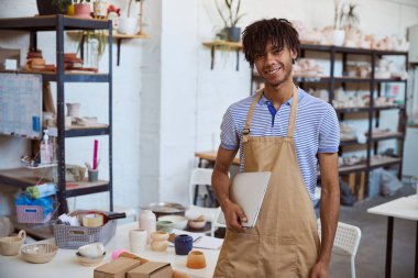 Önlük giyen genç adam çömlek atölyesinde dizüstü bilgisayar tutarken sevinçle gülümsüyor. Gün ışığı, çömlek ve yaratıcı malzemelerle dolu rafları aydınlatıyor..