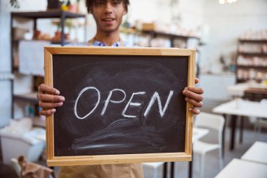 Genç adam elinde açık yazı tahtası tutarken neşeyle gülümsüyor. Arka plandaki parlak perakende alan gündüz vakti hoş bir atmosfer yaratıyor.