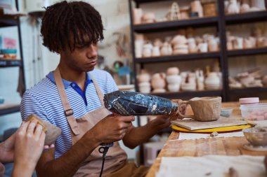 Genç Afrikalı Amerikalı adam çömlek atölyesinde kil nesneler yapmaya odaklanıyor. Isı tabancası ve seramik bardak kullanırken yaratıcılıkla dolu sakin bir atmosfer..