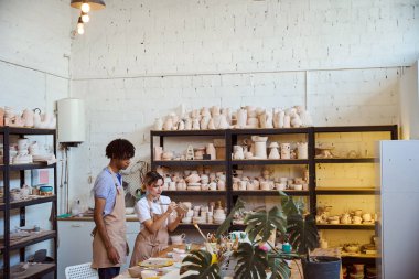 Genç bir kadın çömlek atölyesinde küçük bir seramik nesneyi fırçayla boyamaya odaklanıyor. Bir erkek onun çalışmalarını gözlemler, yaratıcı ve işbirlikçi bir atmosferi ön plana çıkarır..