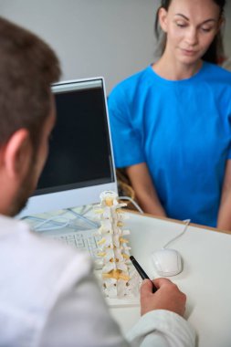 Erkek doktor, odaklanmış bir kadın hastaya detayları açıklarken omurilik modelini işaret ediyor. Doktorun ofisindeki ciddi atmosfer profesyonelliği ve dikkatliliği yansıtıyor..