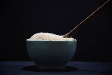 Çiğ beyaz pirinçli porselen tava ve siyah arka planda tahta kaşık. Beyaz pirinç. İnsan yemeği. Nişasta açısından zengin ve besin kaynağı olan tahıllar. Mutfak lezzeti..