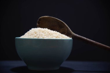 Çiğ beyaz pirinçli porselen tava ve siyah arka planda tahta kaşık. Beyaz pirinç. İnsan yemeği. Nişasta açısından zengin ve besin kaynağı olan tahıllar. Mutfak lezzeti..