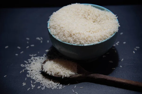 Raw rice served in porcelain frying pan and wooden spoon on dark background. Human food product. Natural food. Cereal grains scattered.