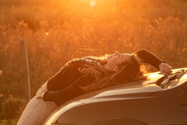 Altın alacakaranlık saatinde bir araçta sırt üstü yatan kadın. Kırsal alan ve alacakaranlıkta dramatik bir gökyüzü. Kırsal fotoğraf sahnesi. Altın saatinde fotoğraf mankeni. Doğal güzellik..