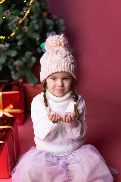 Beautiful little girl in the hat playing with snow near presents and christmas tree. Bokeh lights on the background. Concept christmas — Stock Photo, Image