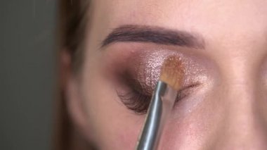 Makyöz, mavi gözlü kadına fırça ve göz farı kullanarak makyaj yapıyor.