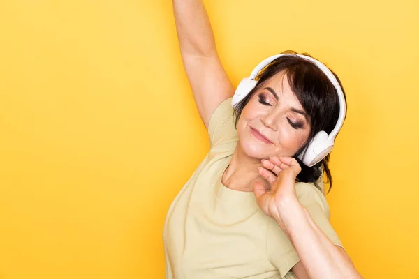 Schöne Seniorin tanzt und entspannt im drahtlosen Kopfhörer vor gelbem Hintergrund — Stockfoto