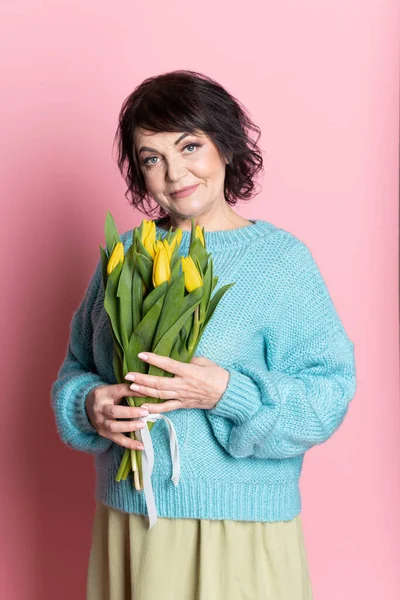 Krásná stará žena s profesionální smokey make-up a účes držení tulipánů květiny na růžovém pozadí — Stock fotografie