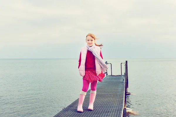 Внешний портрет милой маленькой девочки, играющей у озера в облачный день, в красном платье и розовом жилете, тонированное изображение — стоковое фото